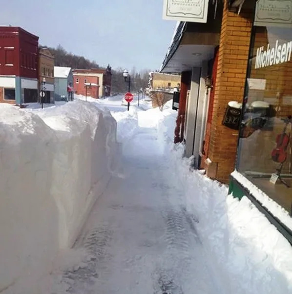 Sidewalk with tall snowbanks, clear path ahead. Storefronts on the right. Quiet town scene