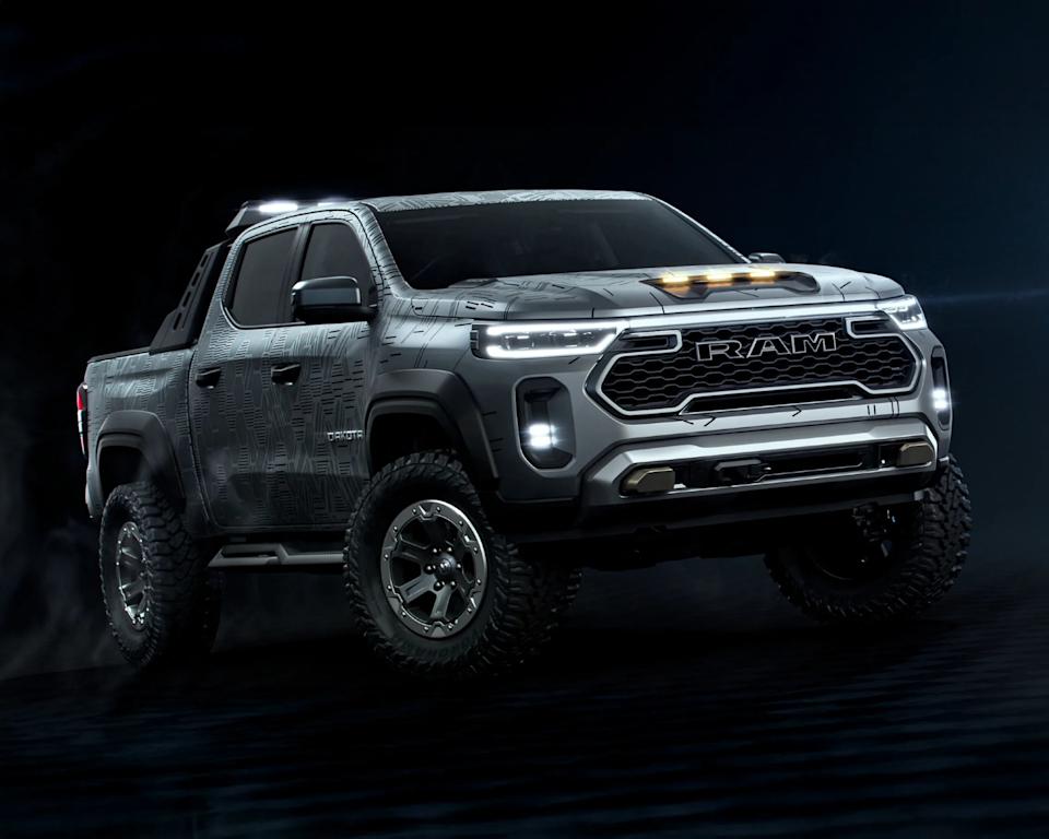 A rugged, gray RAM pickup truck with a bold front grille displaying the RAM logo. The truck features large off-road tires, LED headlights, and additional lights on the hood and front bumper. The body has a textured pattern with the word "Dakota" visible on the side. The truck is shown against a dark background, emphasizing its aggressive and sturdy design.
