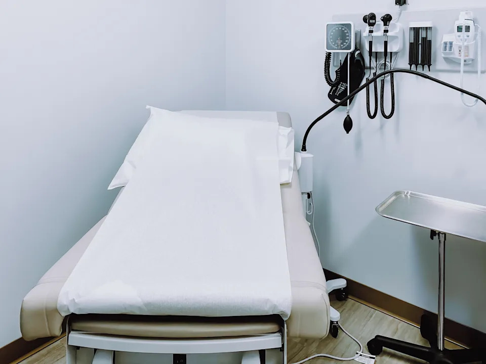 Doctor's office exam room with an empty examination table, medical equipment on the wall, and a tray table nearby