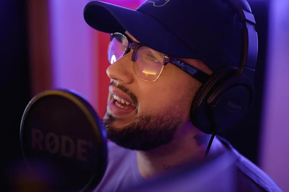 A man with a dark beard, wearing glasses, headphones and a dark ballcap, sings