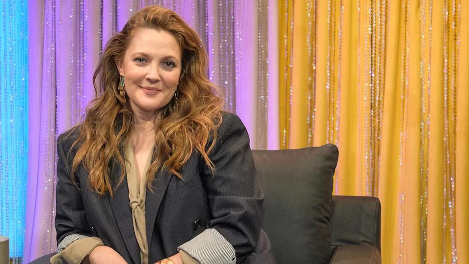 Drew Barrymore con un blazer gris oscuro remangado se sienta en una silla y sonríe suavemente para la cámara