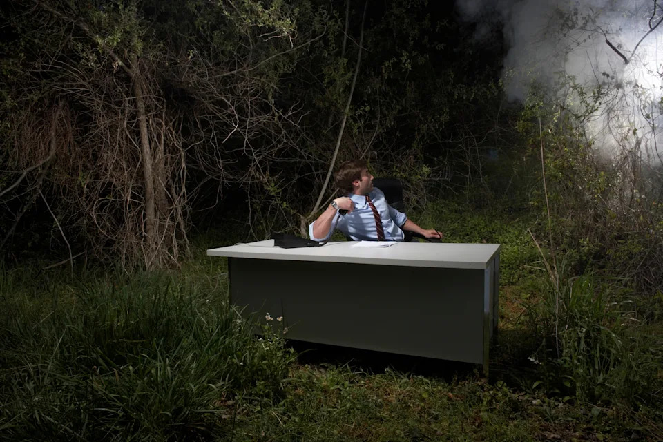 Man in a suit sits at a desk in a forest, looking over his shoulder at smoke in the background