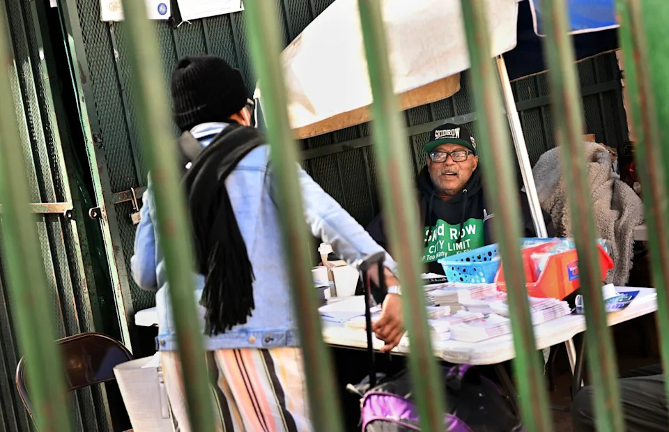A person sits behind a table and talks to a person standing on the other side of the table. Both are behind bars.