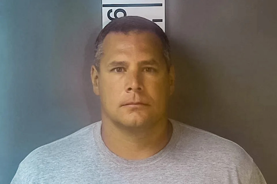 A person stands against a height chart, wearing a plain T-shirt, in a neutral-toned, official setting for a mugshot photo