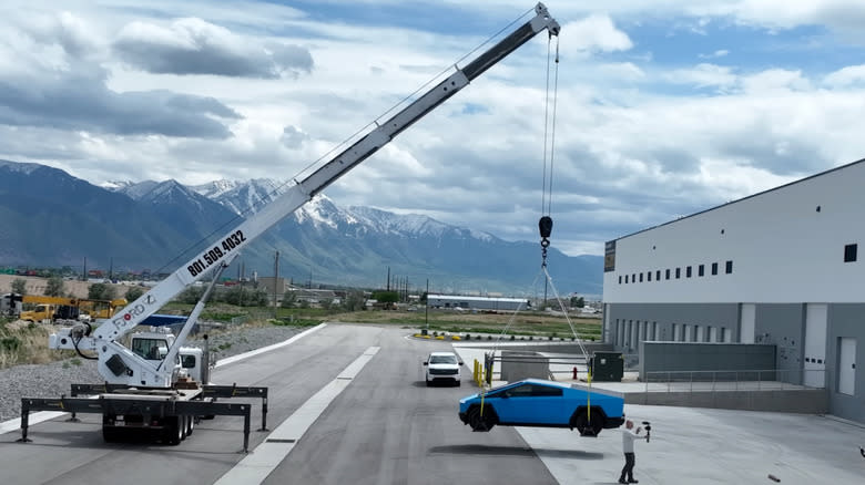 A crane lifts a blue-wrapped Tesla Cybertruck in the air.