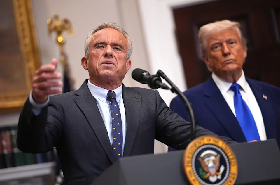 Two men in suits standing at a podium with a presidential seal, one speaking into a microphone. The background includes official decor