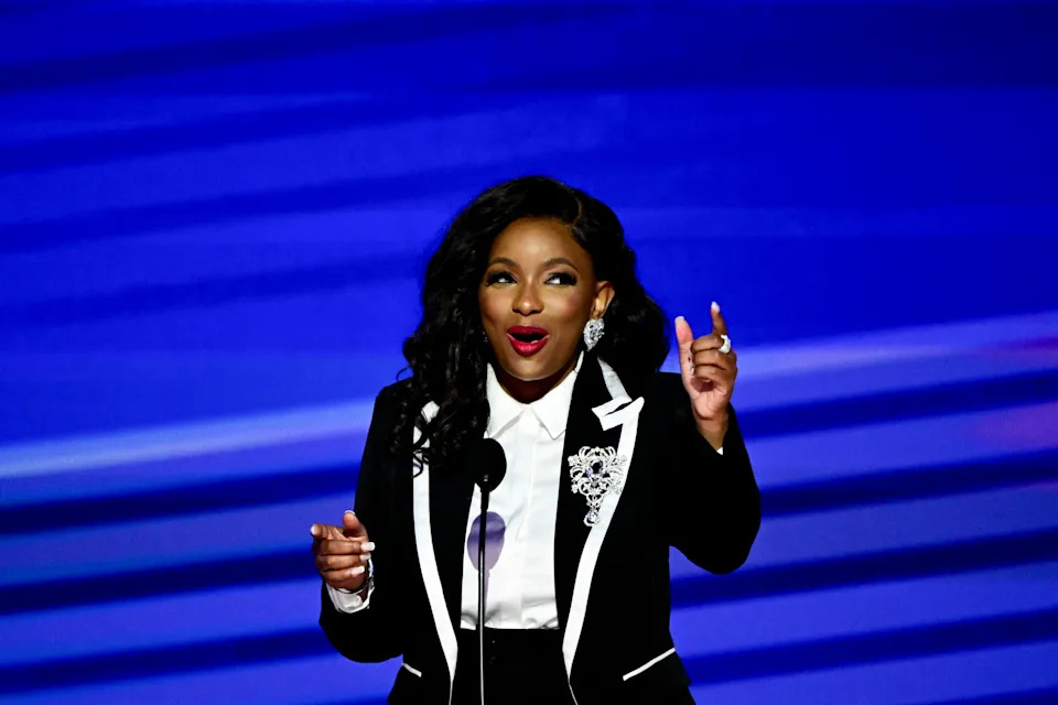 Person in a stylish suit with embellishments speaks animatedly on stage, holding a microphone and gesturing with one hand