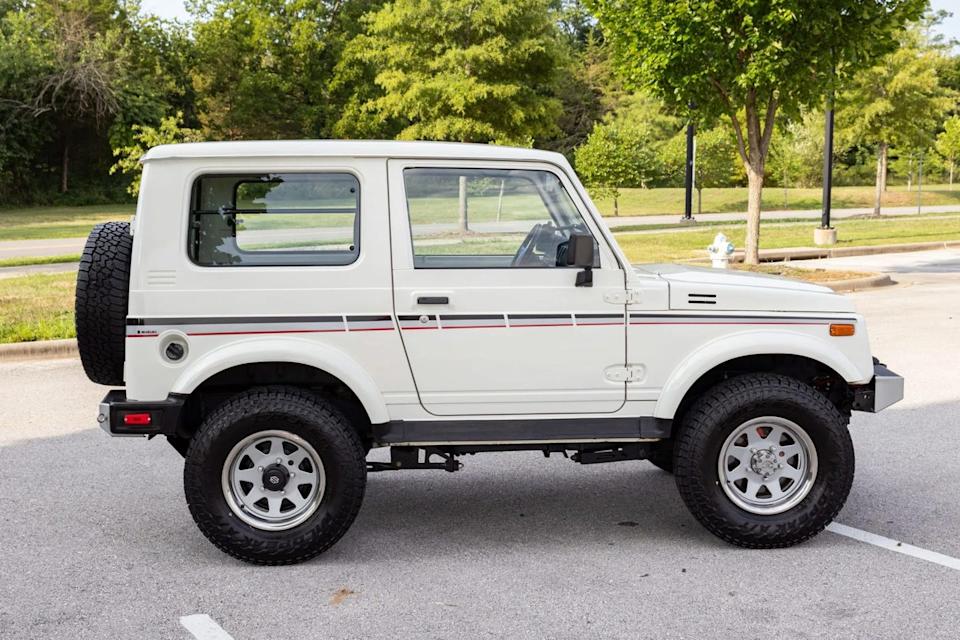 side profile view of white two door suv with black and red stripe parked in lot near trees.