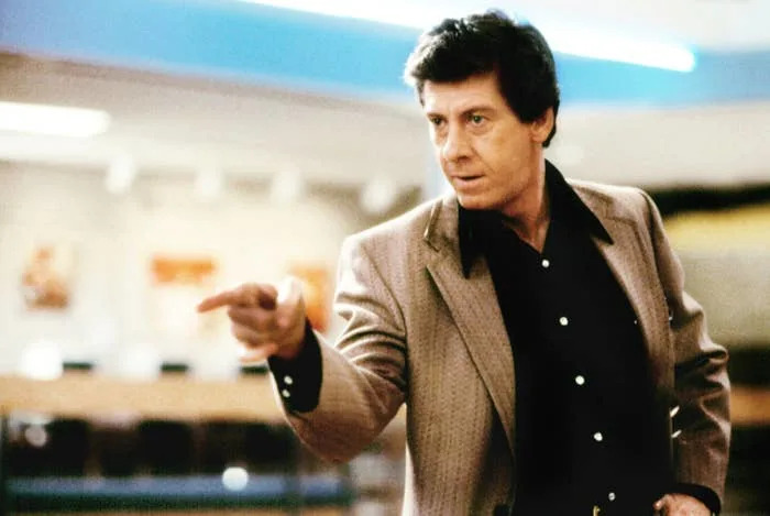 Person in a blazer and shirt points assertively in what appears to be a dramatic scene. Background suggests an indoor setting