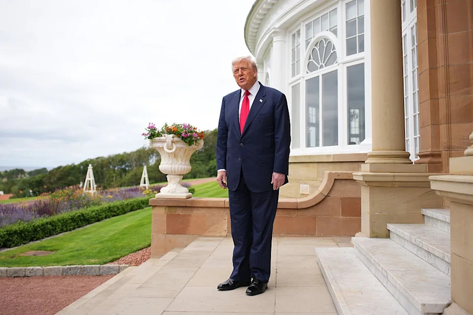 A man in a suit stands on a patio outside a large building, with gardens visible in the background