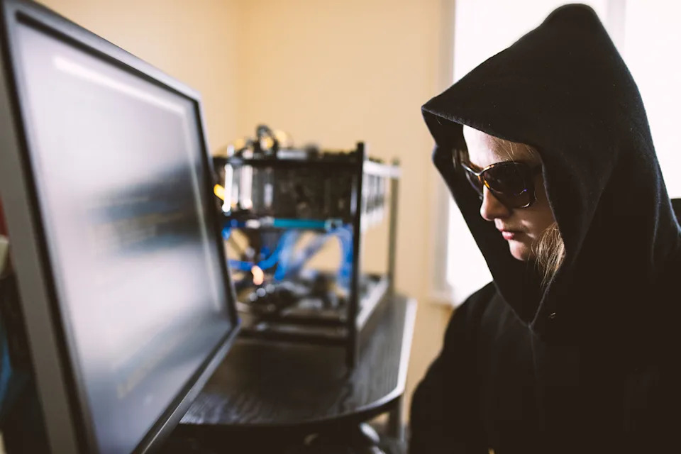 Person in hood and sunglasses at a computer screen, suggesting secrecy or cybersecurity theme