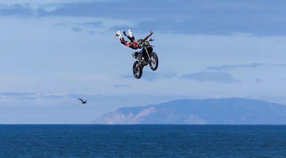 A motorcycle stunt rider extends their arms and legs during a jump, with the ocean visible in the background.