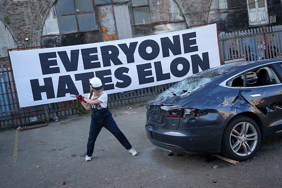 Una mujer golpeando un Tesla con un martillo, con un gran letrero atrás que dice