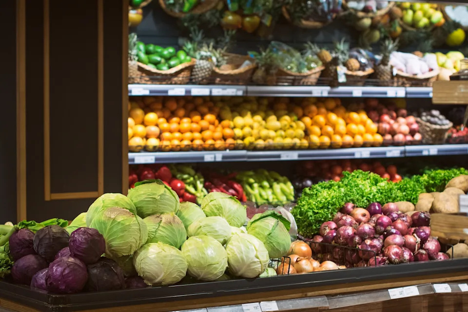 Grocery store produce section with various fruits and vegetables, including cabbages, onions, and citrus fruits