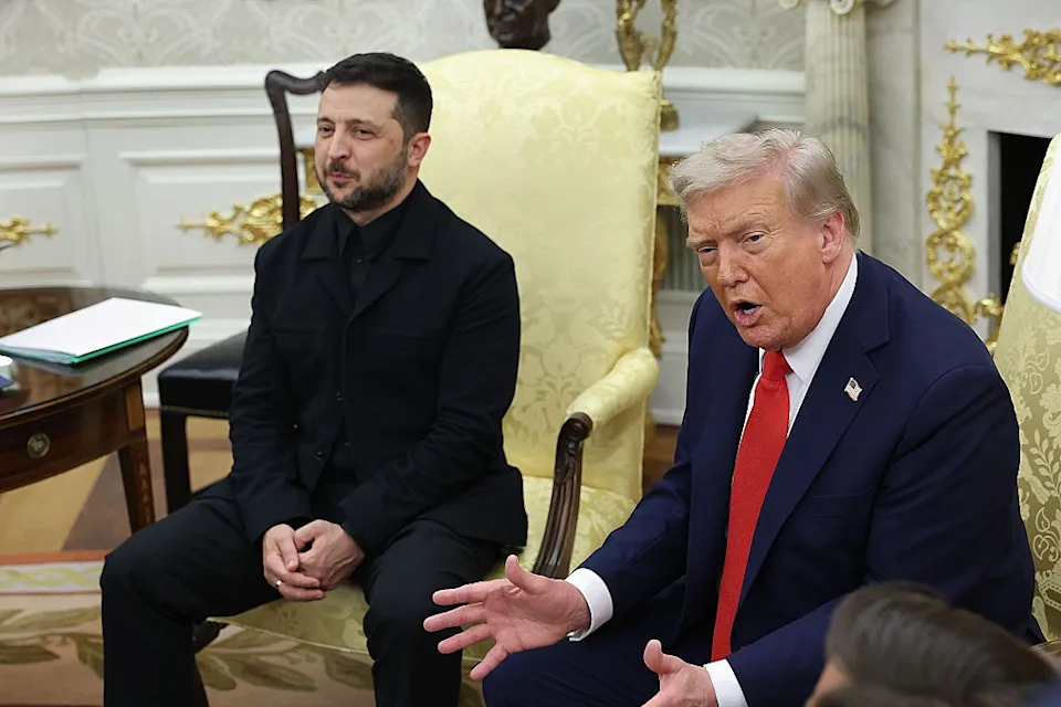 Two people sitting in formal chairs, engaged in discussion. One wears a dark suit, the other a suit with a tie. Elegant setting in the background