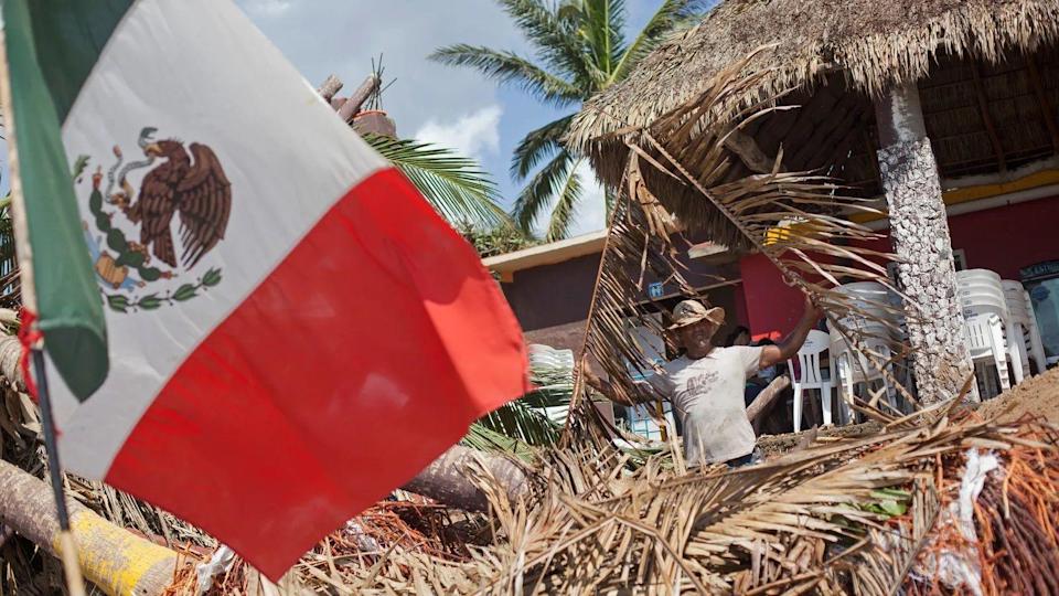 Una imagen muestra la bandera de México en medio de los daños causados por el huracán Patricia. Al fondo se ve un hombre con sombrebro limpiando los escombros.