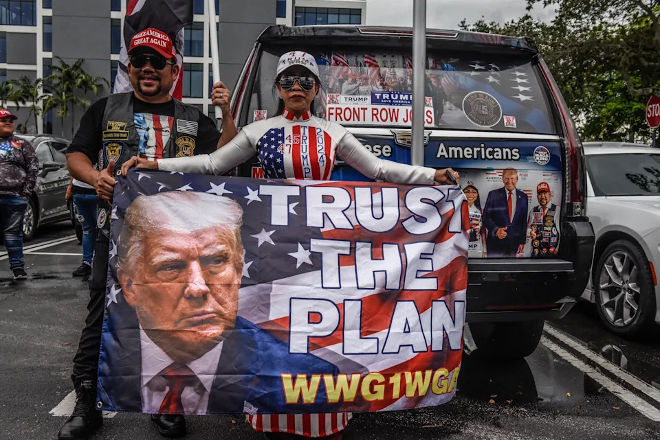 Two individuals stand by a vehicle with pro-Trump posters, holding a large flag featuring Trump's face and the text "Trust the Plan WWG1WGA."