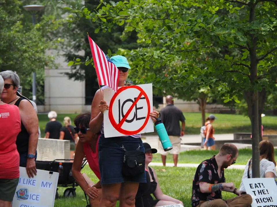 A protester holds an American flag and a sign denouncing U.S. Immigration and Customs Enforcement, or ICE, during a demonstration against President Donald Trump, held beside the Indiana Statehouse on Thursday, July 17, 2025. (Leslie Bonilla Muñiz/Indiana Capital Chronicle)