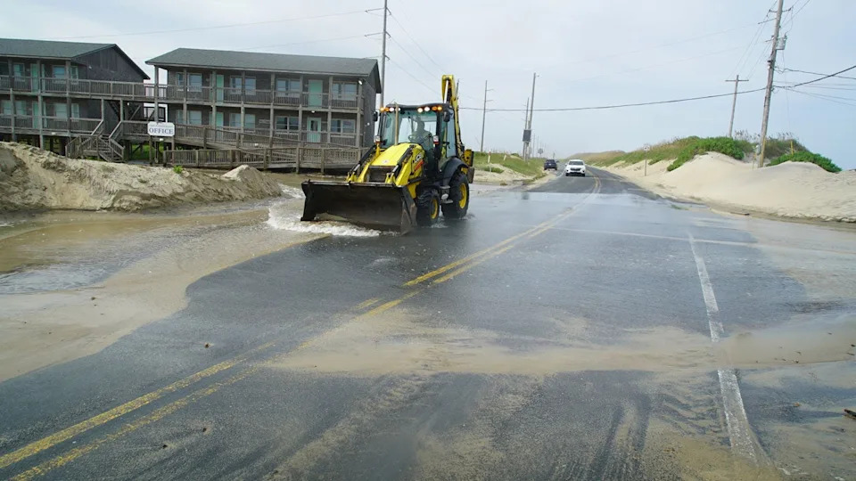 A front-end loader clears a road of sand while floodwaters cover it