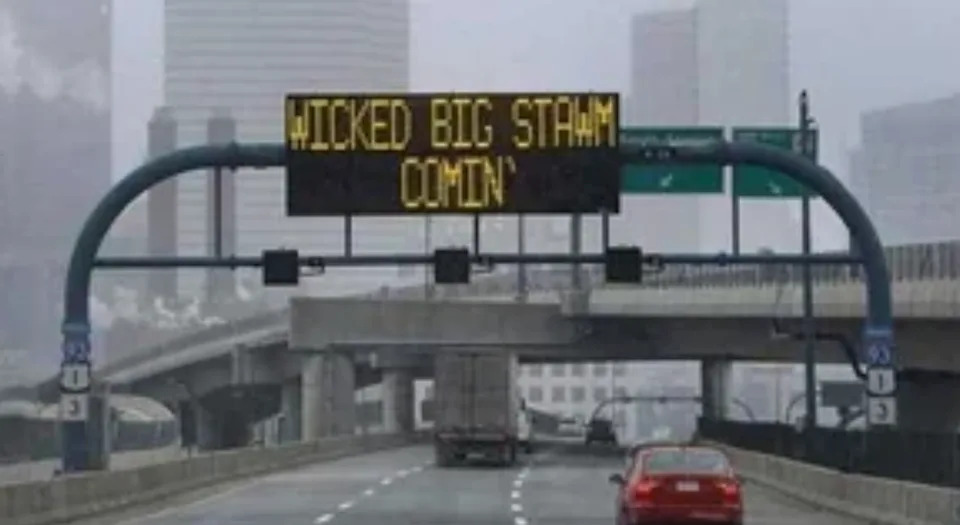 Electronic road sign reads, "Wicked big stahm comin'," warning drivers of an approaching storm in a city setting