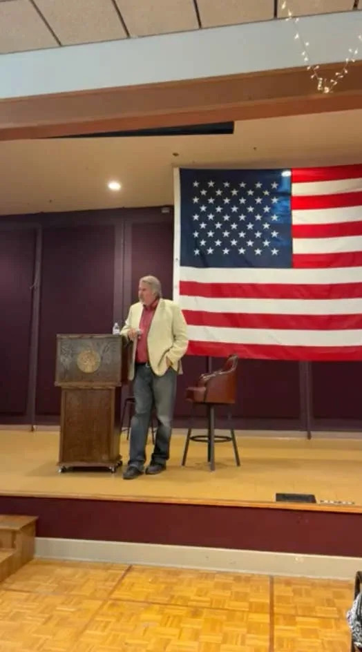 A person in a blazer and jeans speaks at a podium with a large U.S. flag in the background