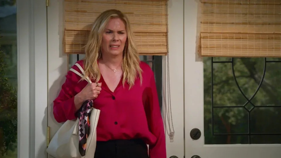 Leanne Morgan stands in front of a door, sweating, wearing a pink shirt and holding a purse