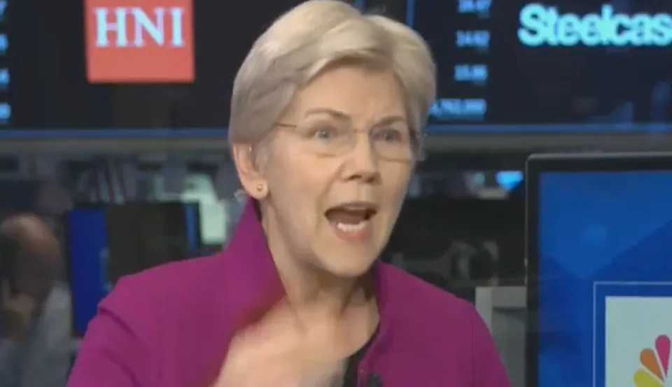 A person speaking passionately during a news interview, wearing a blazer, with a financial news backdrop