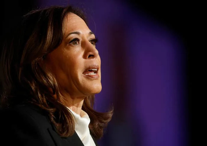 A person speaking at an event, dressed in a dark blazer and white top, likely addressing an audience or media