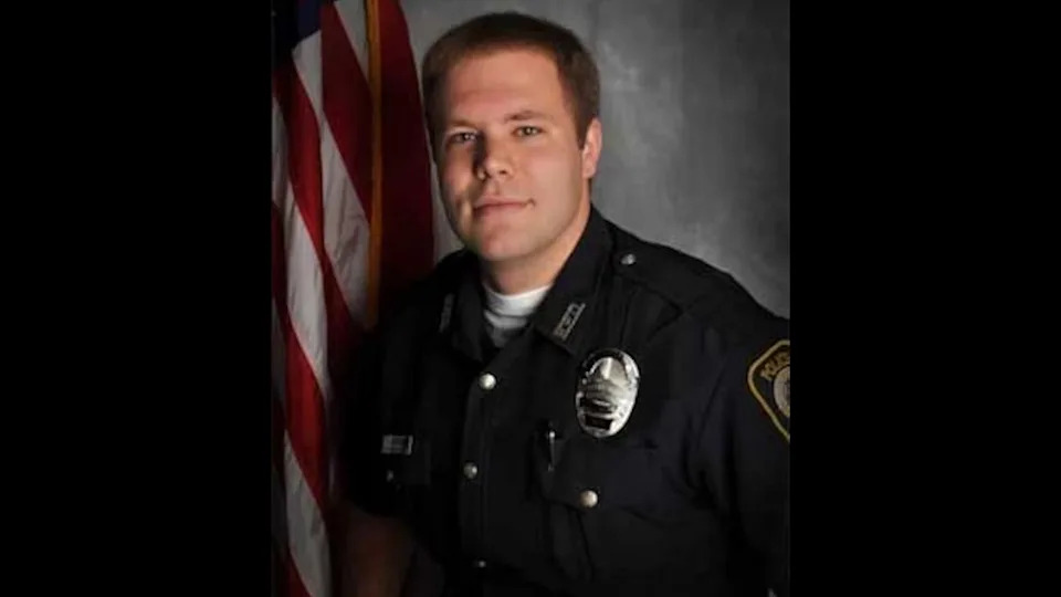 Officer in uniform stands solemnly beside a U.S. flag, gazing at the camera