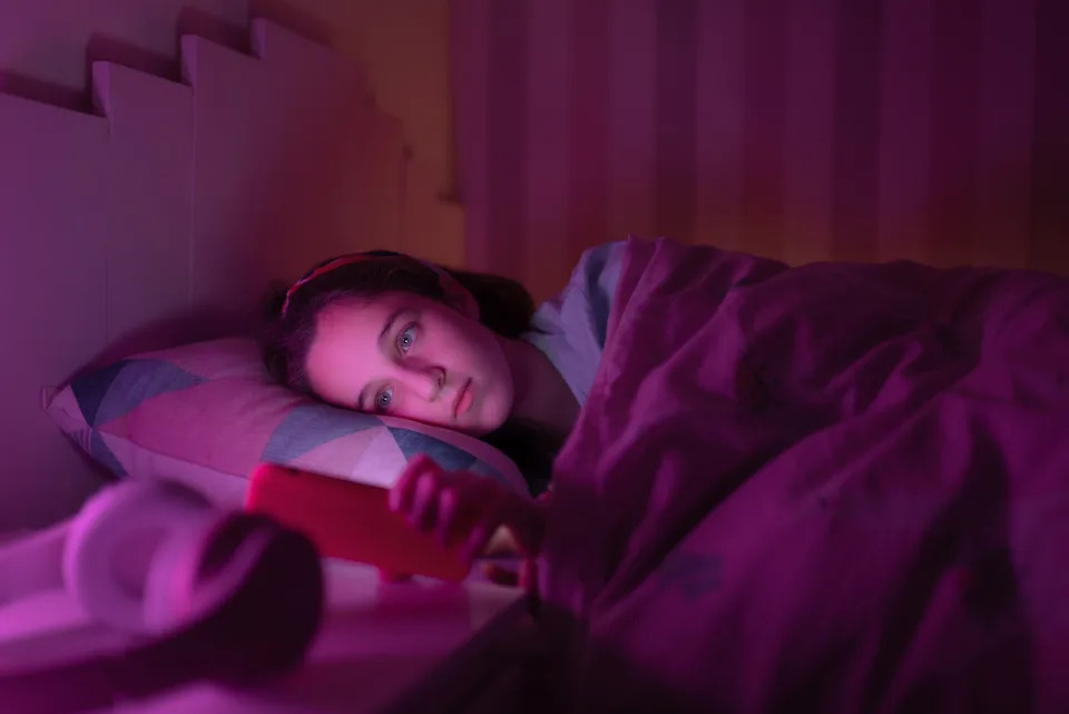 Person lying in bed, looking at their phone with a thoughtful expression, surrounded by a soft ambient light