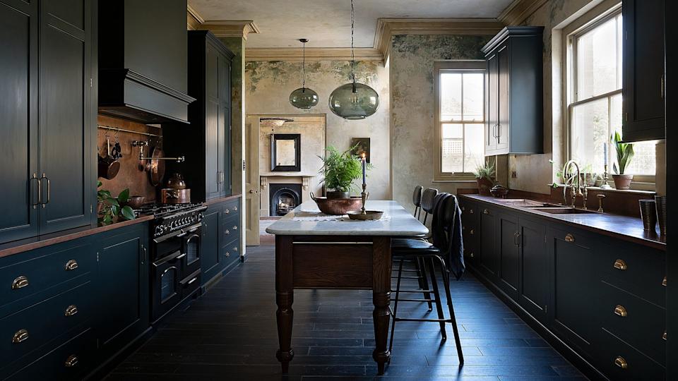Cocina de estilo inglés con los muebles en azul oscuro, el suelo de madera teñida, mesa de madera antigua con el sobre de mármol, taburetes, vela encendida, lámpara de techo de cristal
