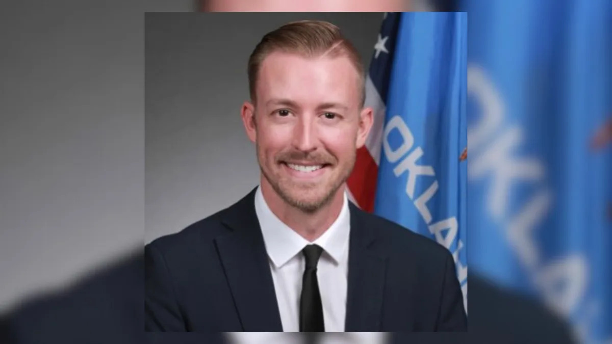 Portrait of a smiling man in a dark suit, white shirt and black tie, standing in front of a blurred background featuring the Oklahoma state flag and the American flag. He has short, neatly styled light brown hair and a trimmed beard. 