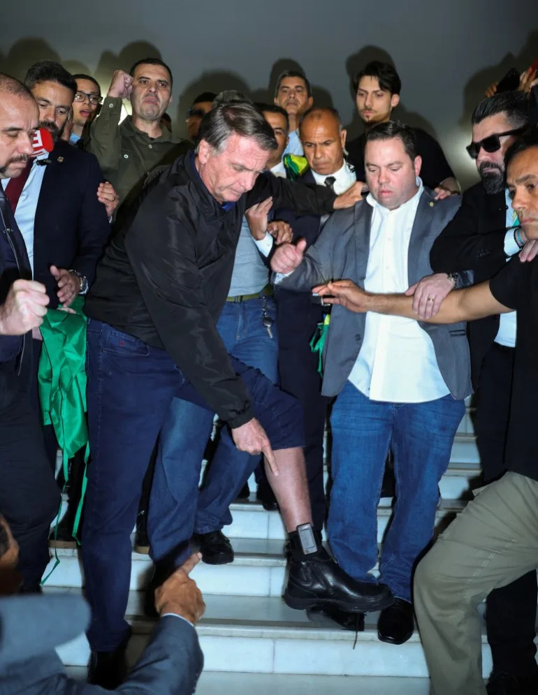 Jair Bolsonaro lifts a pant leg to show his ankle monitor to supporters and news media.