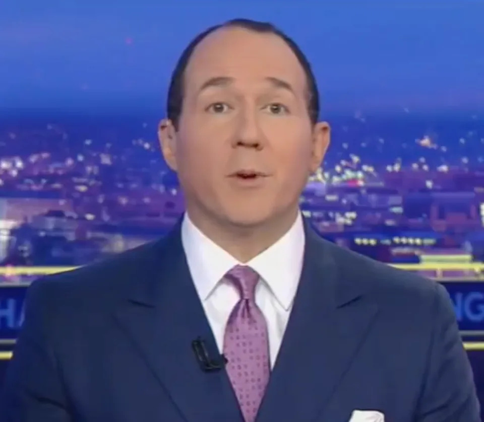 News anchor in a suit and tie delivering a broadcast, cityscape visible in the background