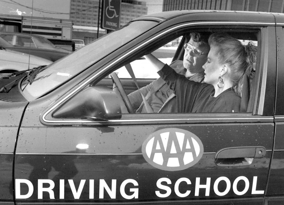 A driving instructor and student in a car labeled "AAA Driving School." The instructor points out of the window, guiding the student