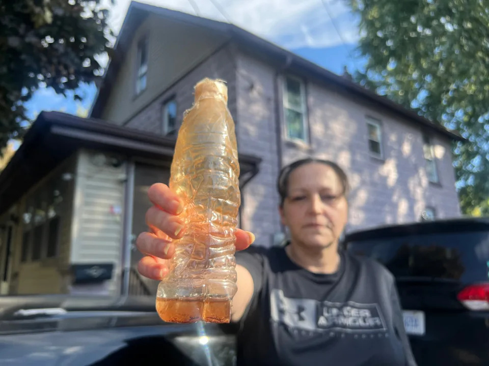 Person holds a plastic bottle filled with murky water, standing in front of a house, highlighting water quality issues