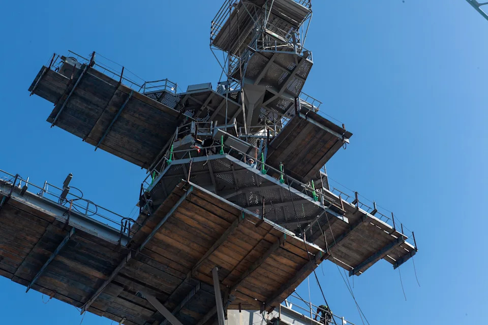 The island of an aircraft carrier is seen from above undergoing maintenance. The sky is clear blue in the background.