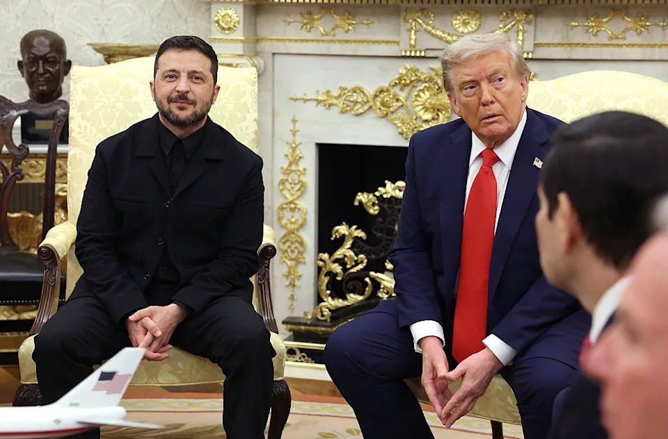 Two men in formal attire sit in an ornate room. One wears a black suit; the other, a blue suit with a red tie. They appear to be in conversation