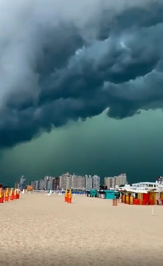 Storm clouds loom over a busy beach with tall buildings in the background, creating an intense and dramatic atmosphere