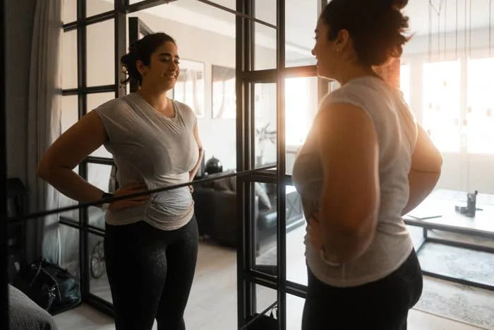 A person smiling at their reflection in a mirror, wearing casual workout clothes, standing with hands on hips in a cozy home setting