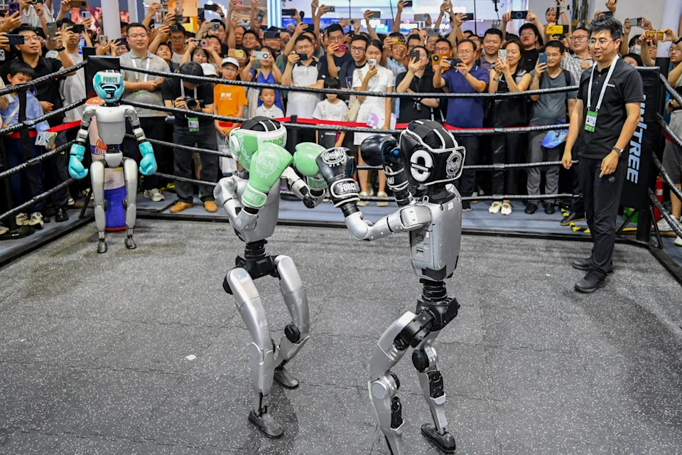 A crowd stands around a boxing ring as two small humanoid robots wearing boxing gloves face off in the center.