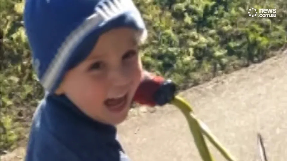Young child with a playful expression, wearing a knit hat, holding onto a tricycle handle outdoors