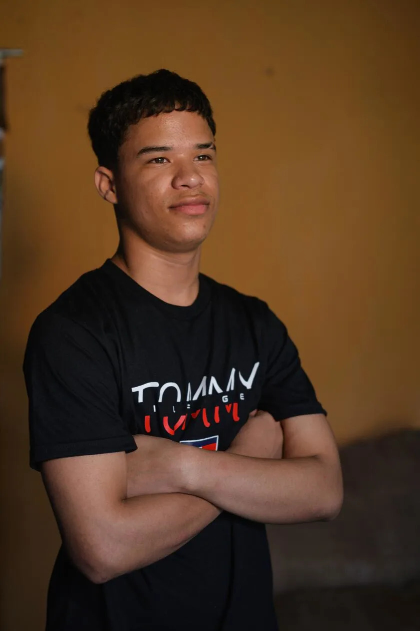 A young man with dark hair, in a dark T-shirt, stands for a portrait with arms crossed