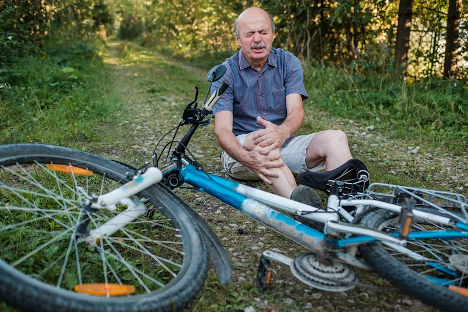 Elderly man in casual attire sits on a path next to a fallen bicycle, holding his knee with a pained expression