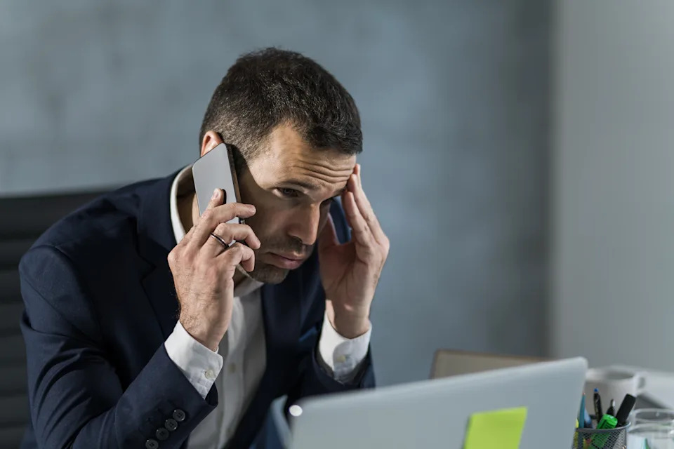 Man in a suit appears stressed, talking on a phone while sitting at a desk with a laptop
