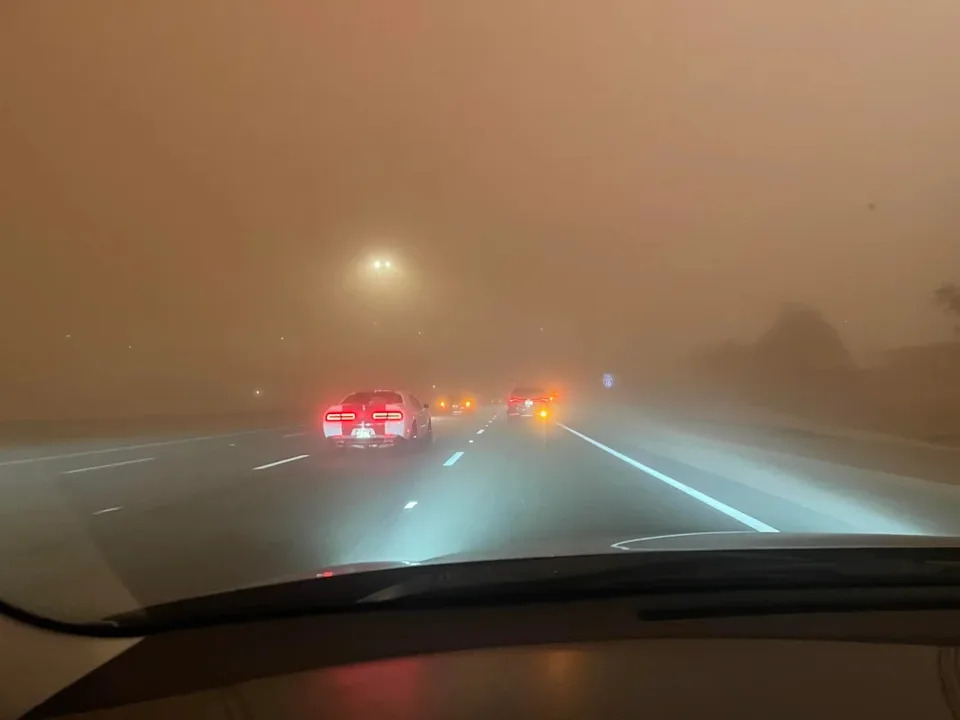 Cars drive on a highway during heavy fog or haze, with visibility greatly reduced. Headlights and taillights are on, illuminating the blurry scene