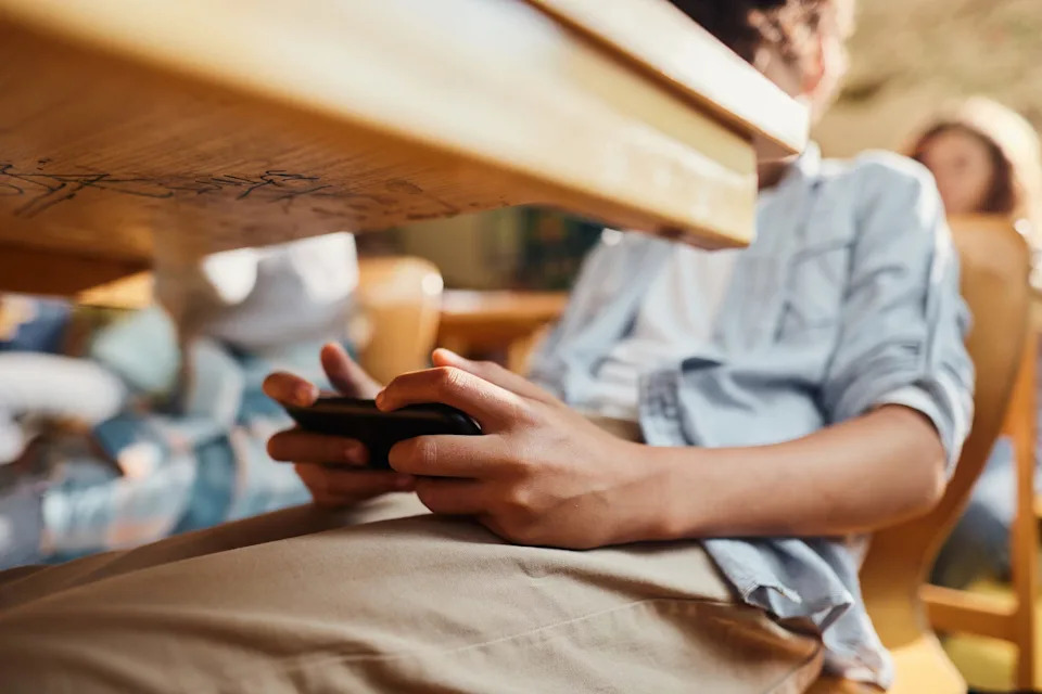 Close up of unrecognizable student using cell phone under the table during a class at elementary school.