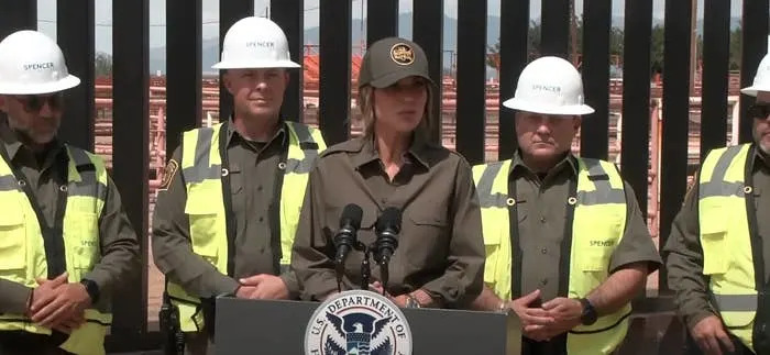 A person in a uniform and hat stands at a podium with "U.S. Department of" seal, flanked by officials in vests and helmets
