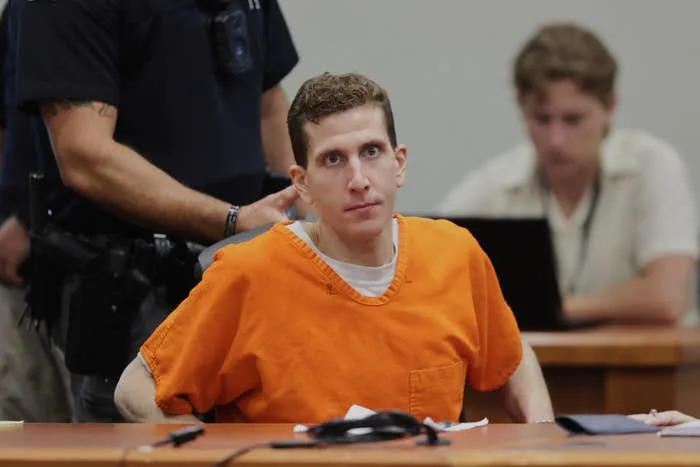 A man in orange prison attire sits in court with a serious expression, while others work in the background