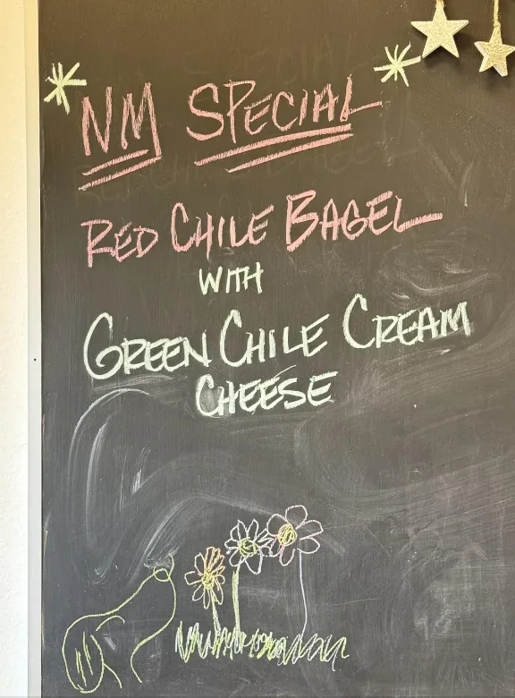 Chalkboard sign reading: "NM Special - Red Chile Bagel with Green Chile Cream Cheese" with doodles of flowers and a snail